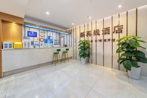 Área de recepção de hotel com balcão, cadeiras, relógio, cartazes informativos e plantas em vasos.