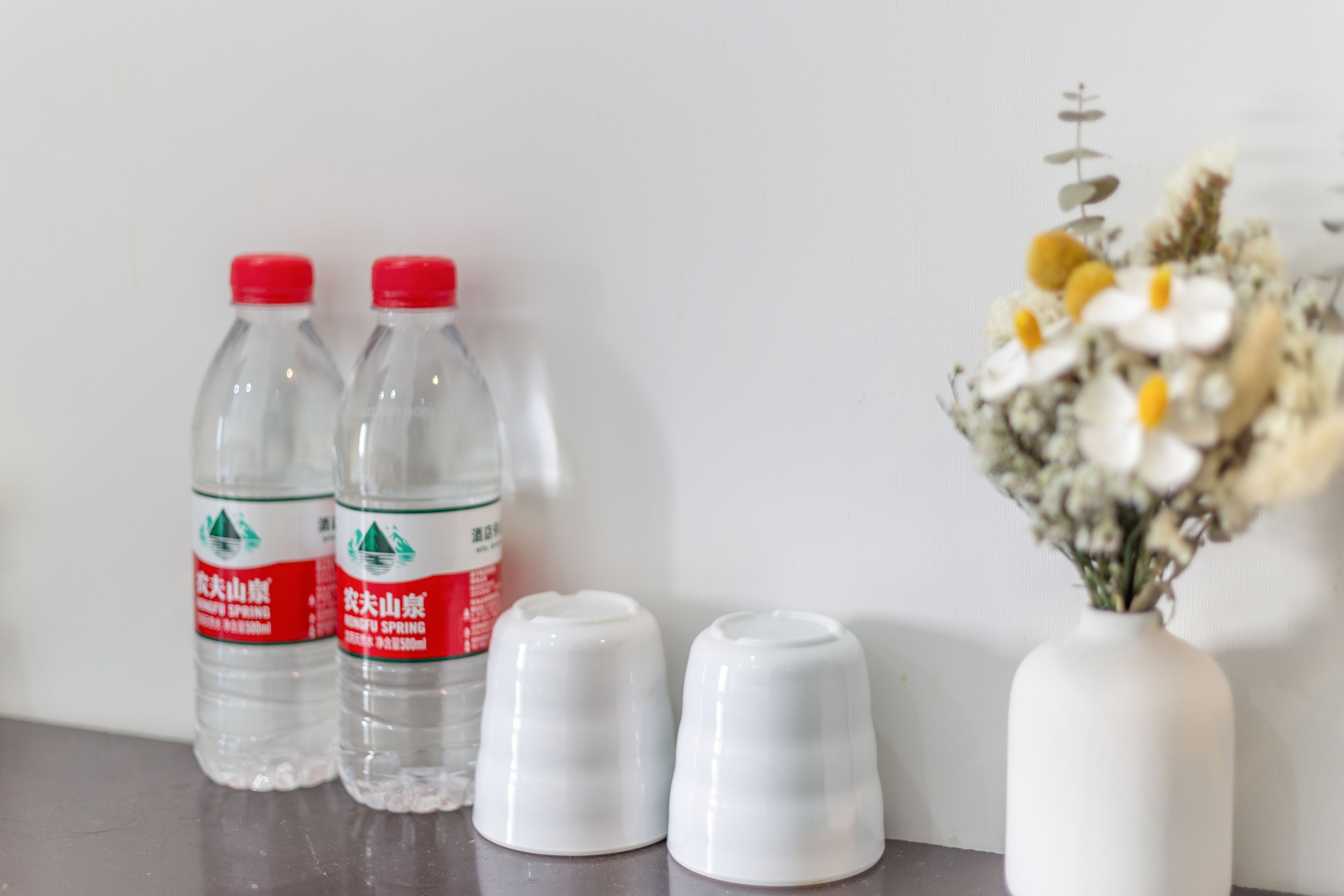 Two clear water bottles with red caps and a white vase with flowers on a table.