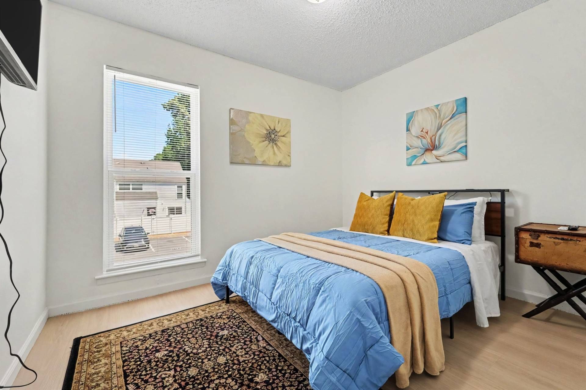 A bedroom with a bed, a window with blinds, and two wall-mounted paintings.