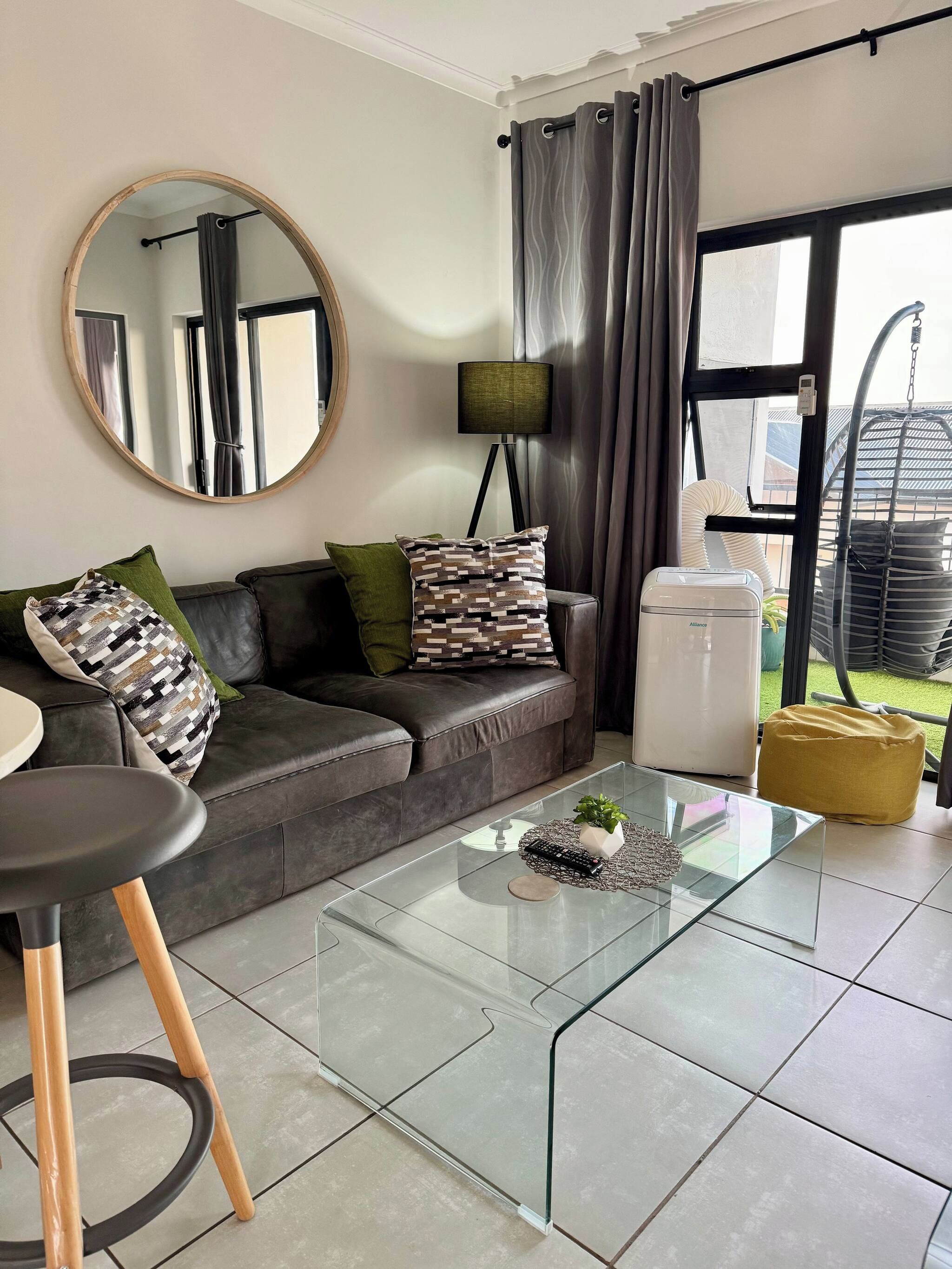 Uma sala de estar moderna com uma mesa de centro de vidro, um sofá com almofadas estampadas, um espelho redondo, um abajur e vista para o exterior através de uma grande janela.