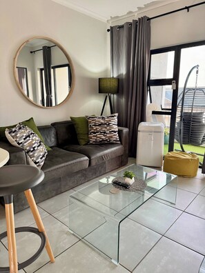 A modern living room with a glass coffee table, a sofa with patterned cushions, a round mirror, a floor lamp, and a view of the outdoors through a large window.
