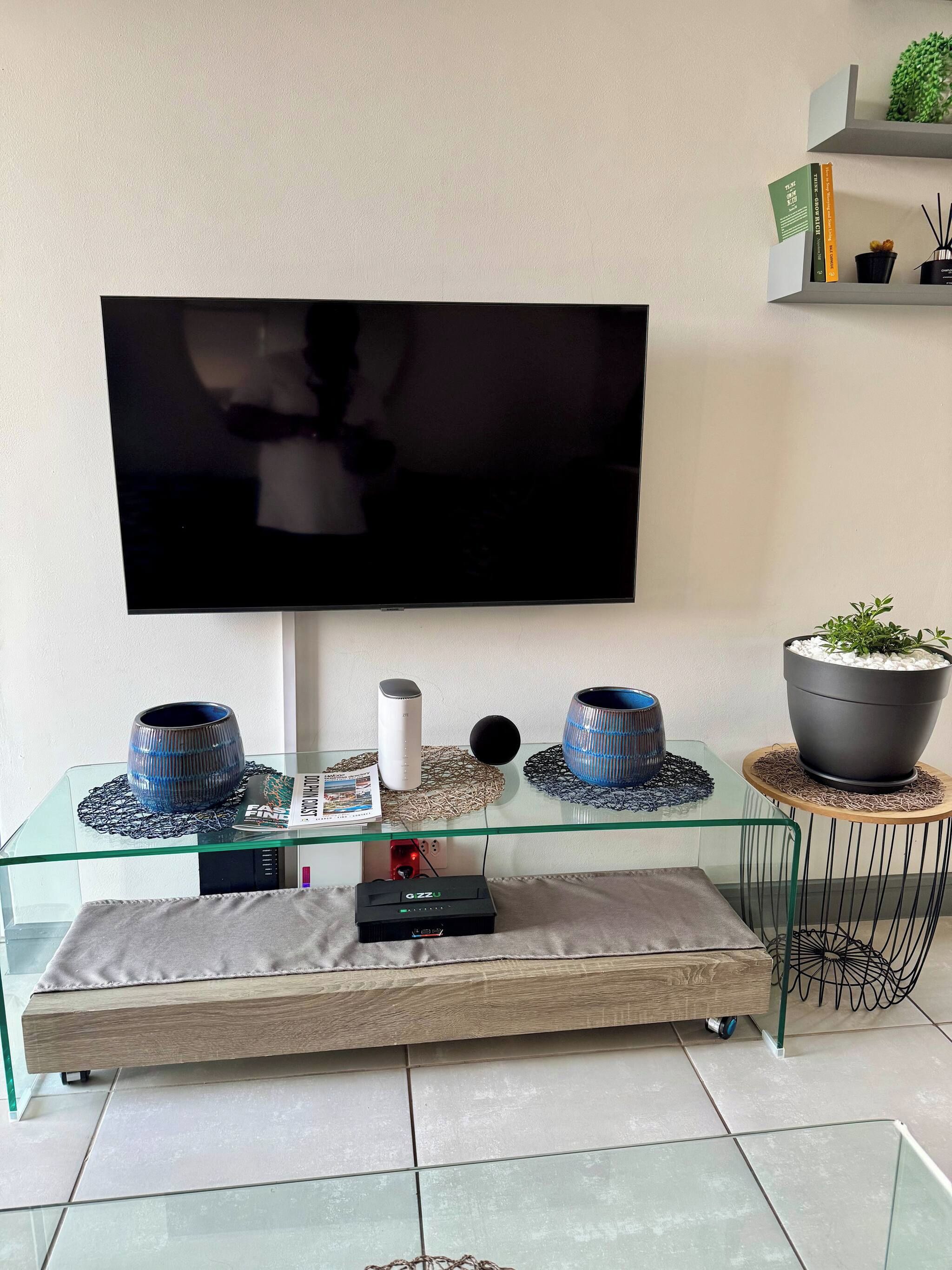 A modern living room with a flat-screen TV mounted on the wall, a glass coffee table with decorative items, a sofa, and a small side table with a potted plant.