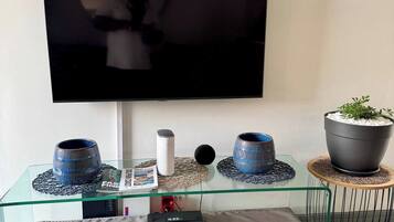 A modern living room with a flat-screen TV mounted on the wall, a glass coffee table with decorative items, a sofa, and a small side table with a potted plant.
