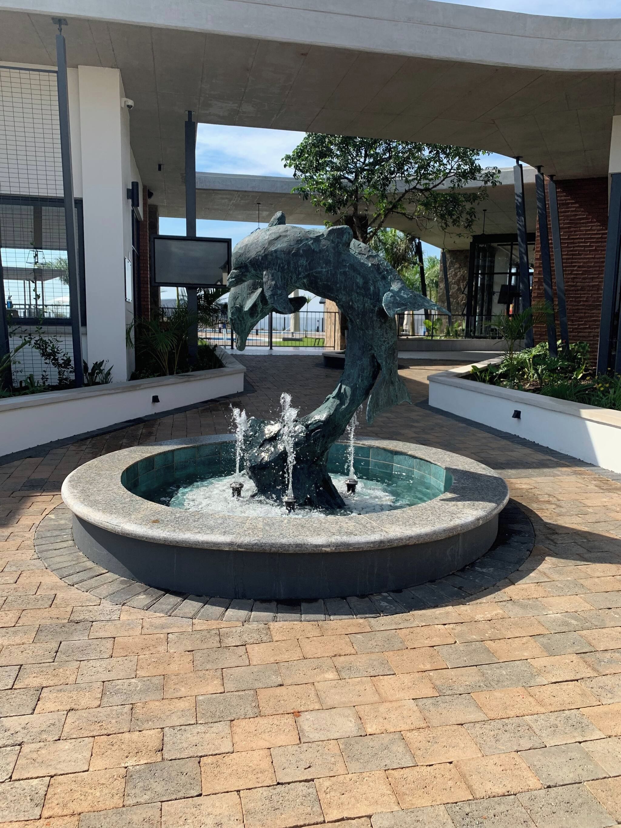 A fountain with a dolphin sculpture in the center, surrounded by a paved area with a building in the background.
