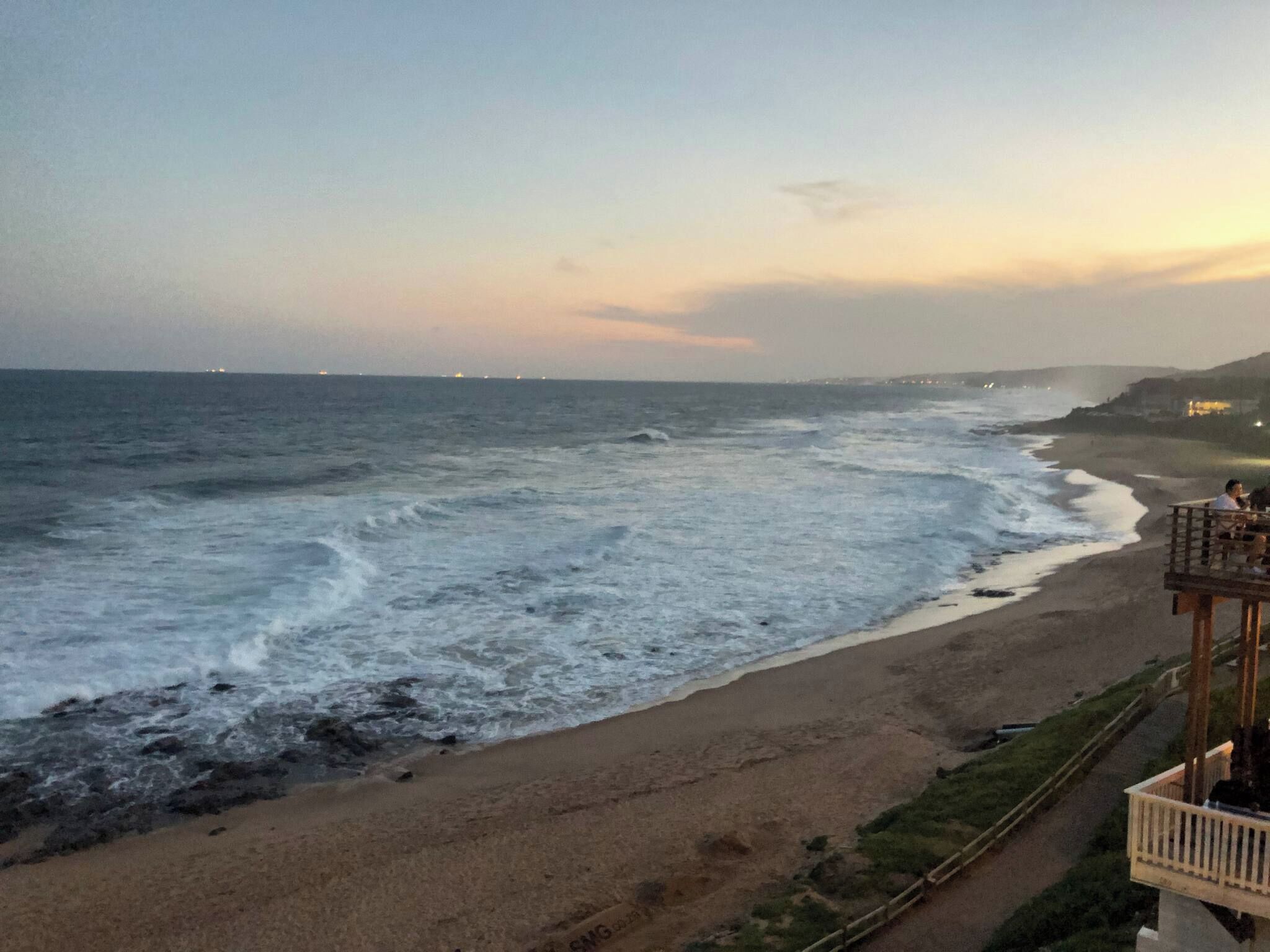 Uma praia com ondas, areal e um edifício com varanda com vista para o mar.