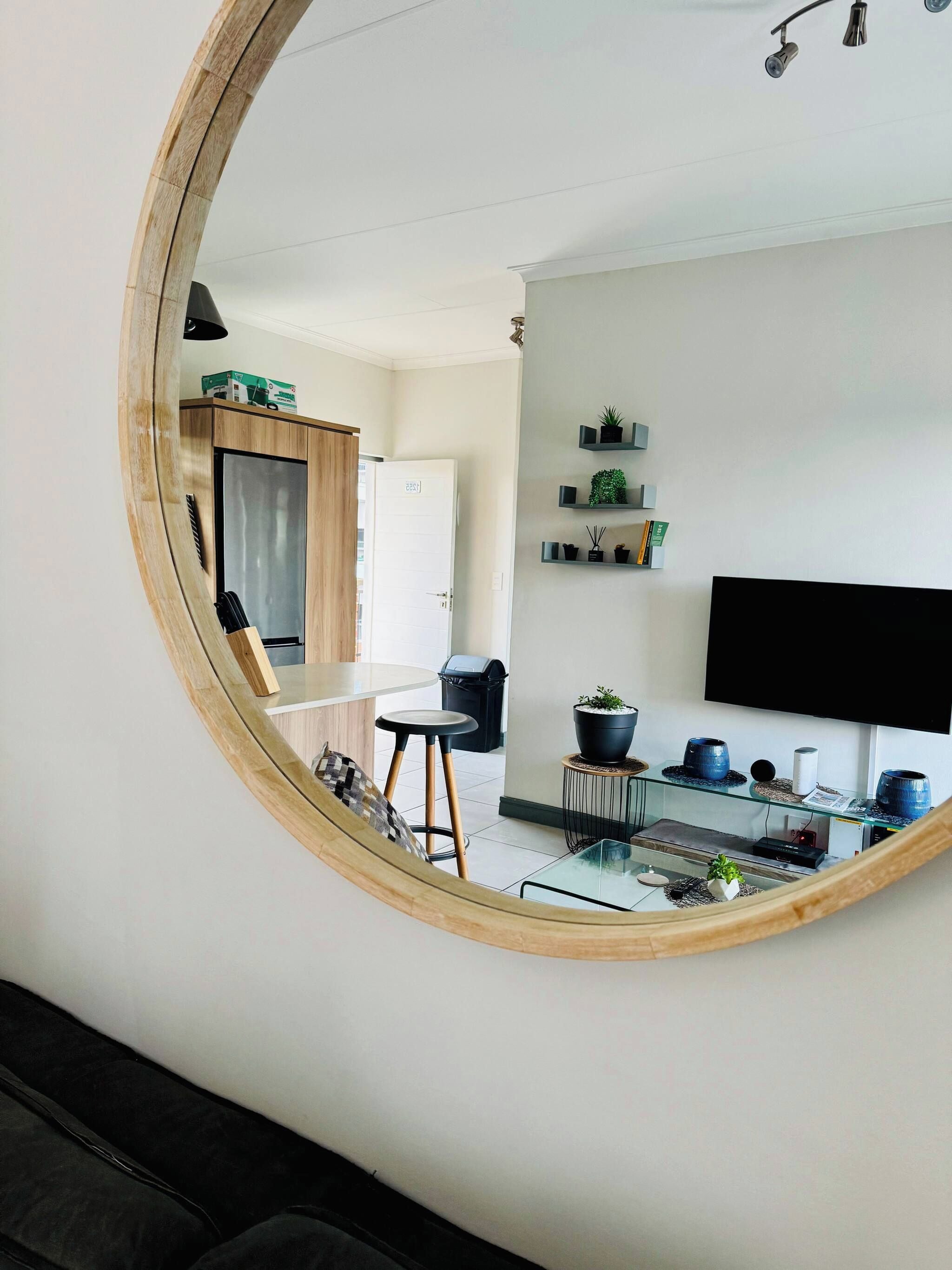 A modern living room with a large mirror reflecting the space, a flat-screen TV mounted on the wall, a glass coffee table, and a small side table with decorative items.