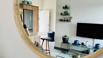 A modern living room with a large mirror reflecting the space, a flat-screen TV mounted on the wall, a glass coffee table, and a small side table with decorative items.