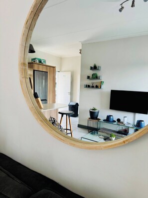 Una sala de estar moderna con un espejo grande que refleja el espacio, un televisor de pantalla plana montado en la pared, una mesa de centro de vidrio y una mesita auxiliar con objetos decorativos.