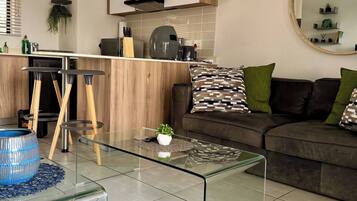 A modern living room with a glass coffee table, a sofa with patterned cushions, and a kitchen area with wooden cabinets and a hanging plant.