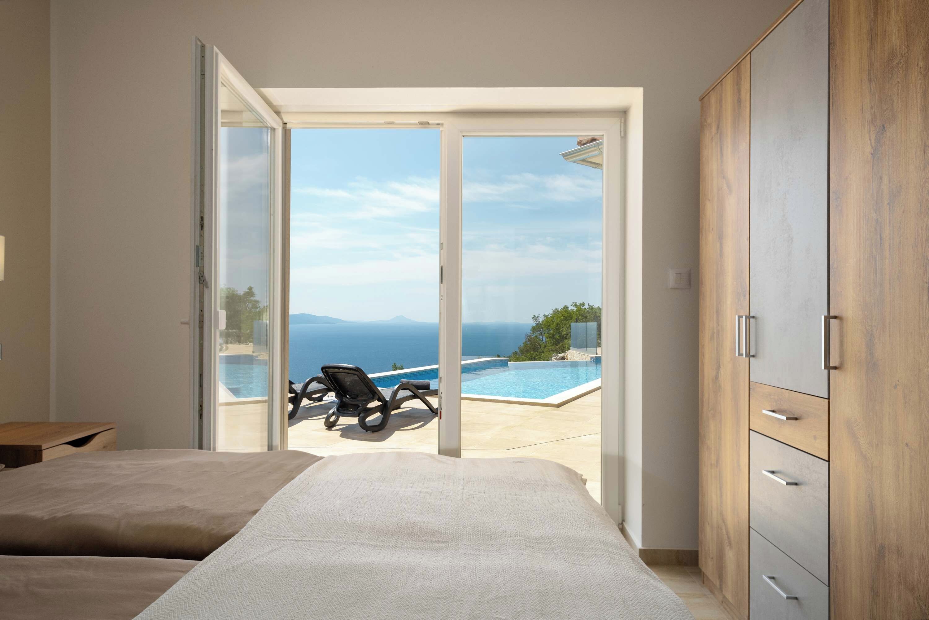 Une chambre avec un lit, une armoire et une porte-fenêtre coulissante donnant sur un espace piscine avec vue sur la mer.