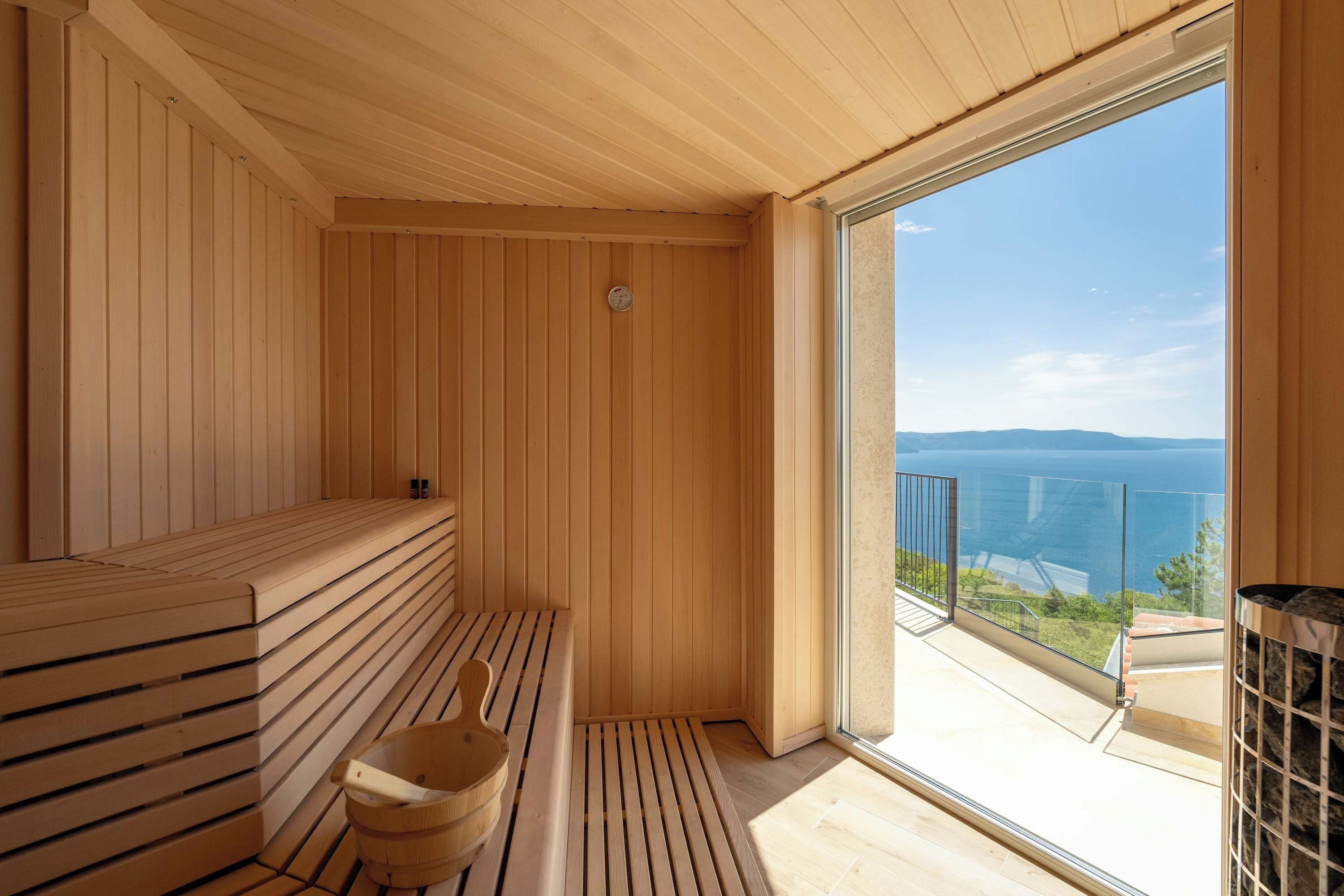 Una sauna con panche in legno e un catino di legno, una porta vetrata che dà su un balcone con vista mare.