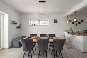 A modern dining area with a wooden table, grey upholstered chairs, and a kitchen area with a white countertop and hanging pendant lights. - Villa Mare Visum with heated pool (Ripenda Kosi)