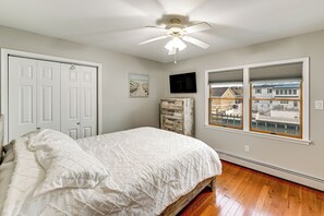 Un dormitorio con cama, ventilador de techo, televisor y ventana con vistas a edificios.