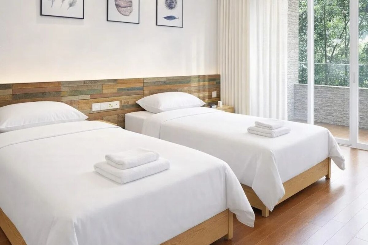 A hotel room with two beds, a wooden headboard, and a view of greenery through large windows.