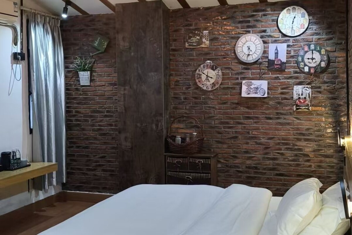 A bedroom with a large bed, wooden beams on the ceiling, and a brick wall with clocks and pictures.