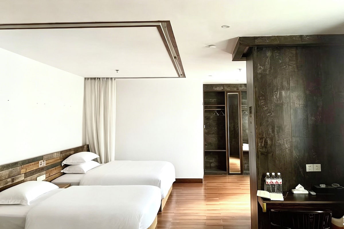 A hotel room with two beds, a wooden headboard, a dark accent wall, a desk with a chair, and a wardrobe.