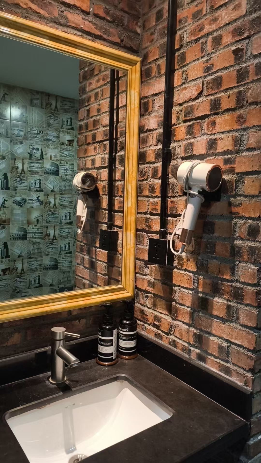 A bathroom with a brick wall, a large mirror, a sink, and two hair dryers mounted on the wall.