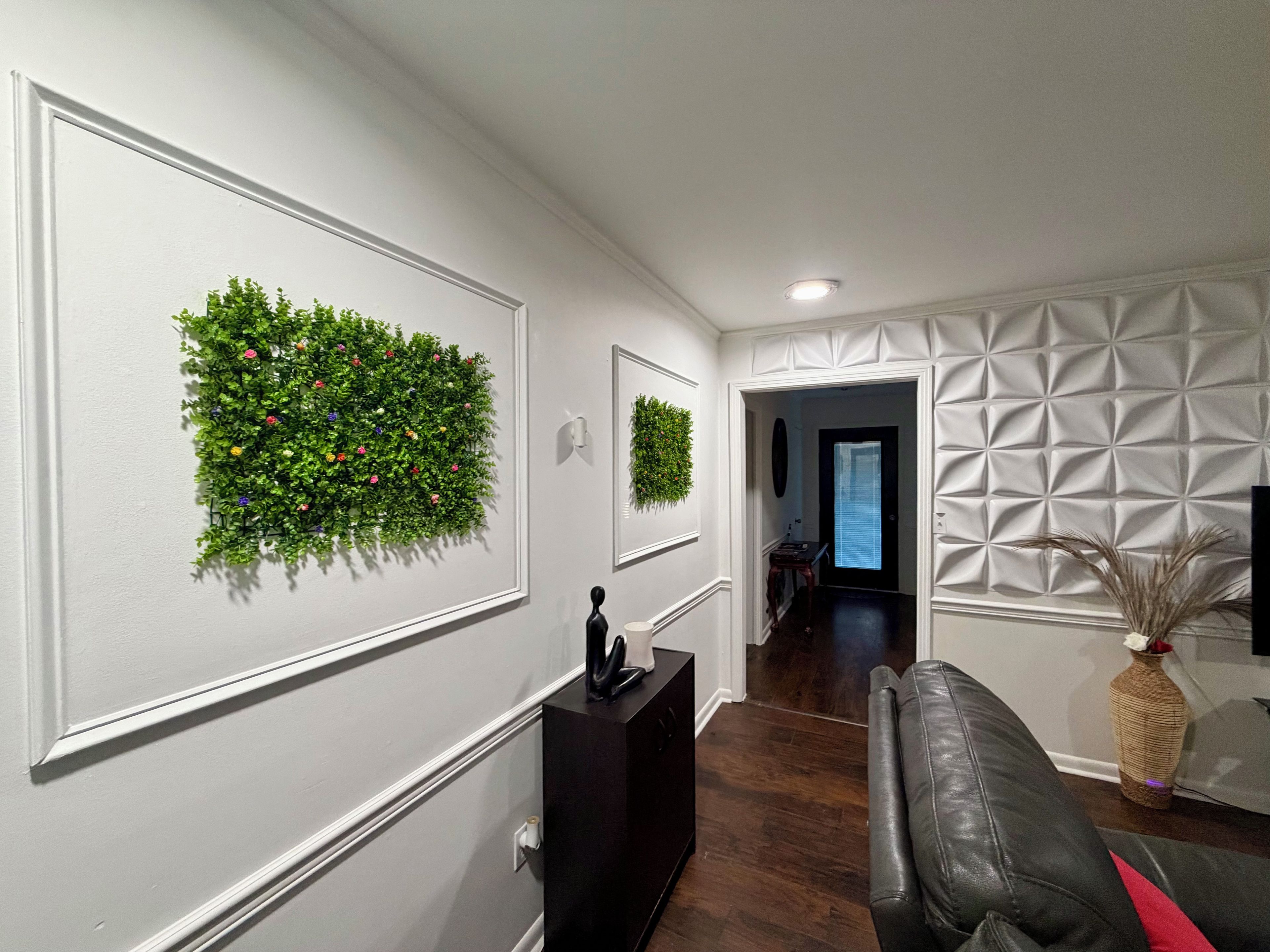 Un couloir avec un parquet en bois foncé, des murs blancs et un canapé en cuir noir. On y trouve deux jardinières murales ornées de plantes vertes et un miroir décoratif.