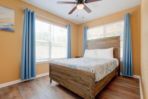 Ein Schlafzimmer mit einem hölzernen Bett, einem Deckenventilator und großen Fenstern mit blauen Vorhängen.