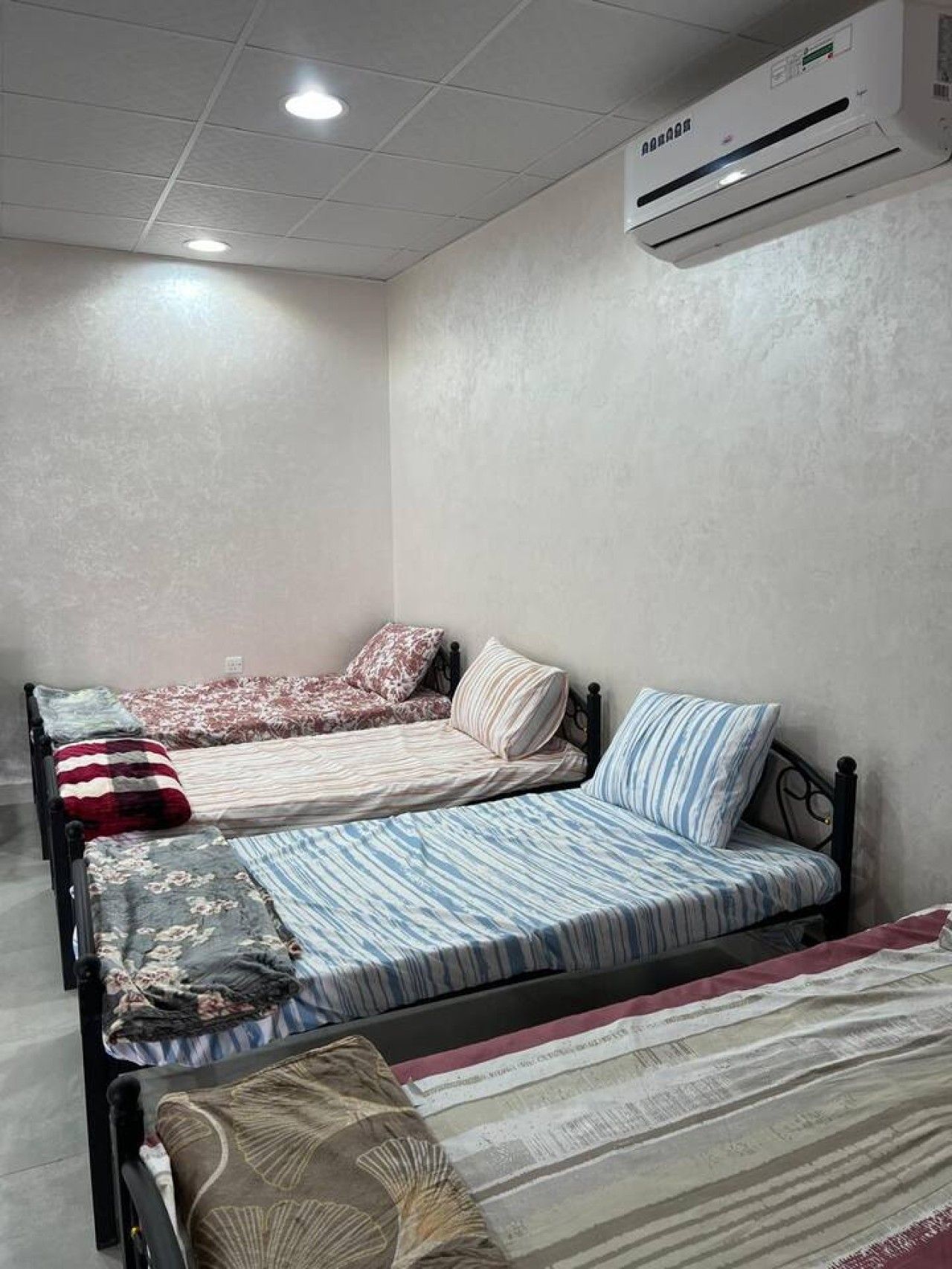 Habitación con tres camas, cada una con diferentes diseños de ropa de cama. Se observa un equipo de aire acondicionado instalado en la pared.