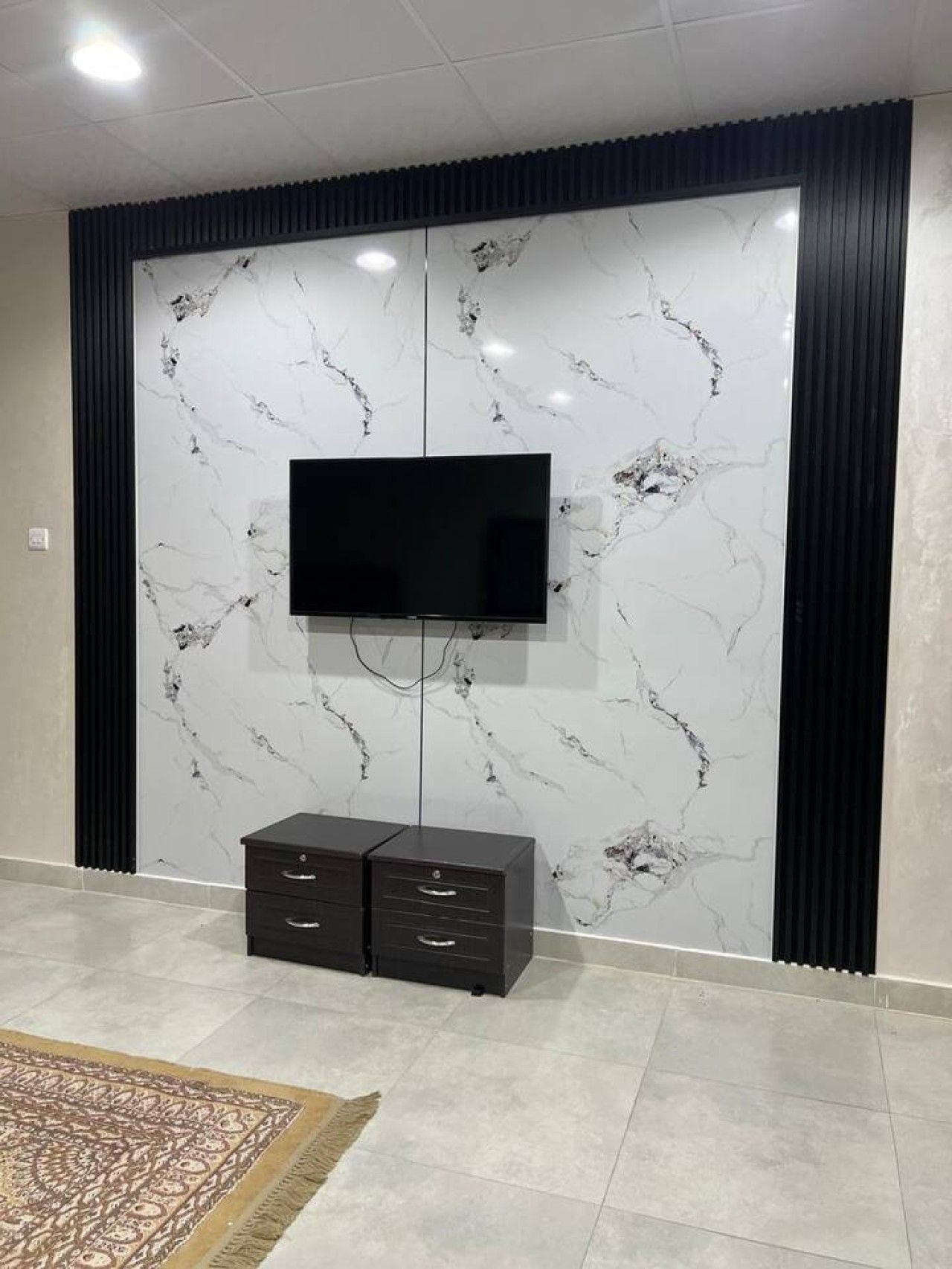 Una habitación con un televisor montado en una pared de mármol, un mueble negro en la parte inferior y una alfombra estampada en el suelo.