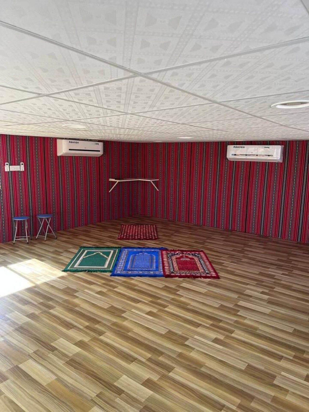 Una habitación con alfombra roja estampada, suelo de madera, dos unidades de aire acondicionado y tres esterillas para orar.