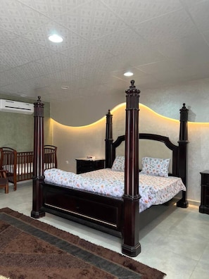 Una cama con dosel con una colcha estampada, una mesita de noche de madera, una silla y un equipo de aire acondicionado de pared.