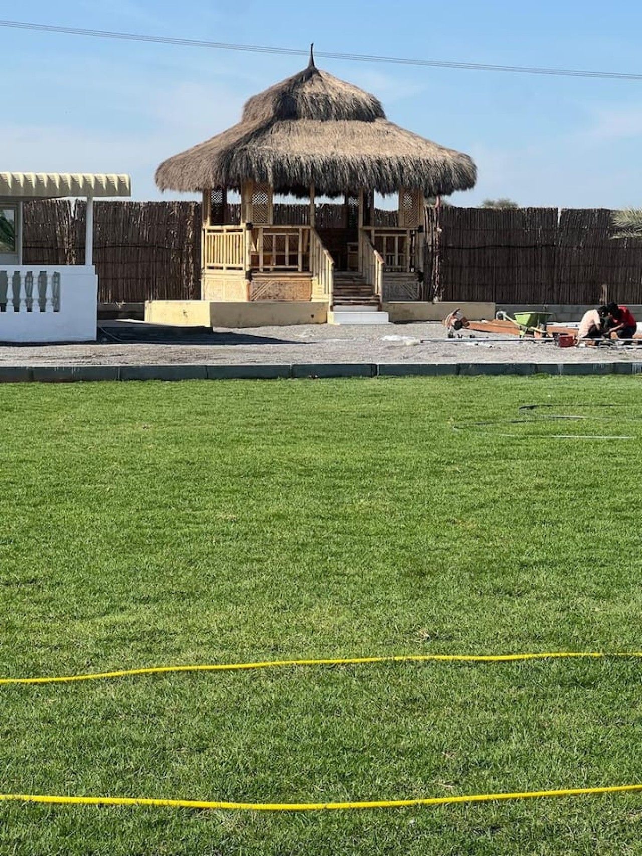 A thatched-roof gazebo with a staircase leading to a platform, surrounded by a wooden fence and a grassy area with yellow lines.