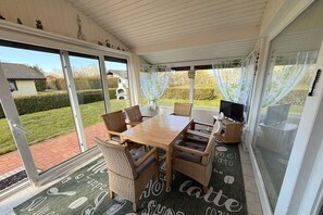 Een overdekt terras met een houten tafel, rieten stoelen, een televisie en uitzicht op de tuin.