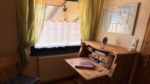 Habitació petita amb escriptori de fusta, cadira, radiador, finestra amb cortines i un quadre encimbellat a la paret.