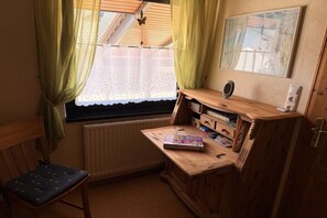 Une petite pièce avec un bureau en bois, une chaise, un radiateur, une fenêtre avec des rideaux et un tableau encadré accroché au mur.