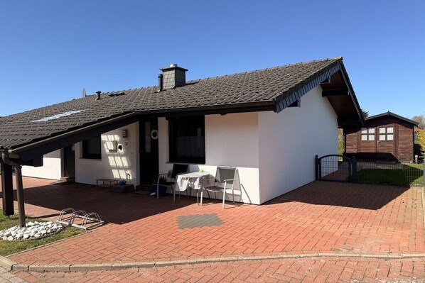 Een gelijkvloers huis met een schuin dak, een terras met tafel en stoelen, en een kleine tuin.