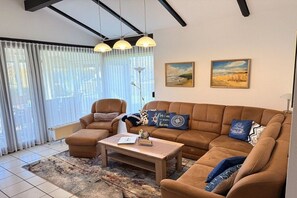 Un salon avec un canapé marron, une table basse et deux fauteuils. Il y a deux tableaux au mur, un lampadaire et une fenêtre avec des rideaux légers.