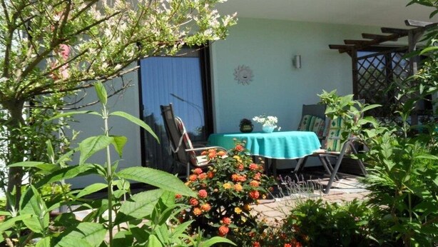 Zona de terrassa amb taula i cadires, envoltada de vegetació i plantes florejants.