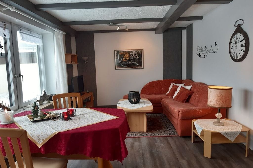 A cozy living room with a red sofa, a dining table with a red tablecloth, and a wall clock.