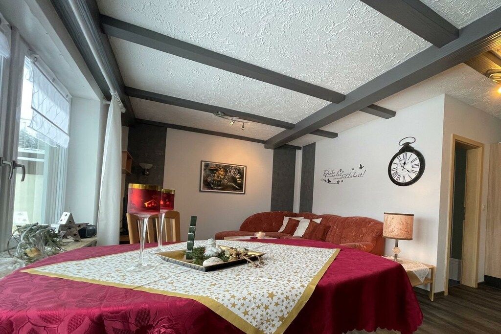 A room with a bed covered in a red blanket, a wall clock, and a decorative wall piece.