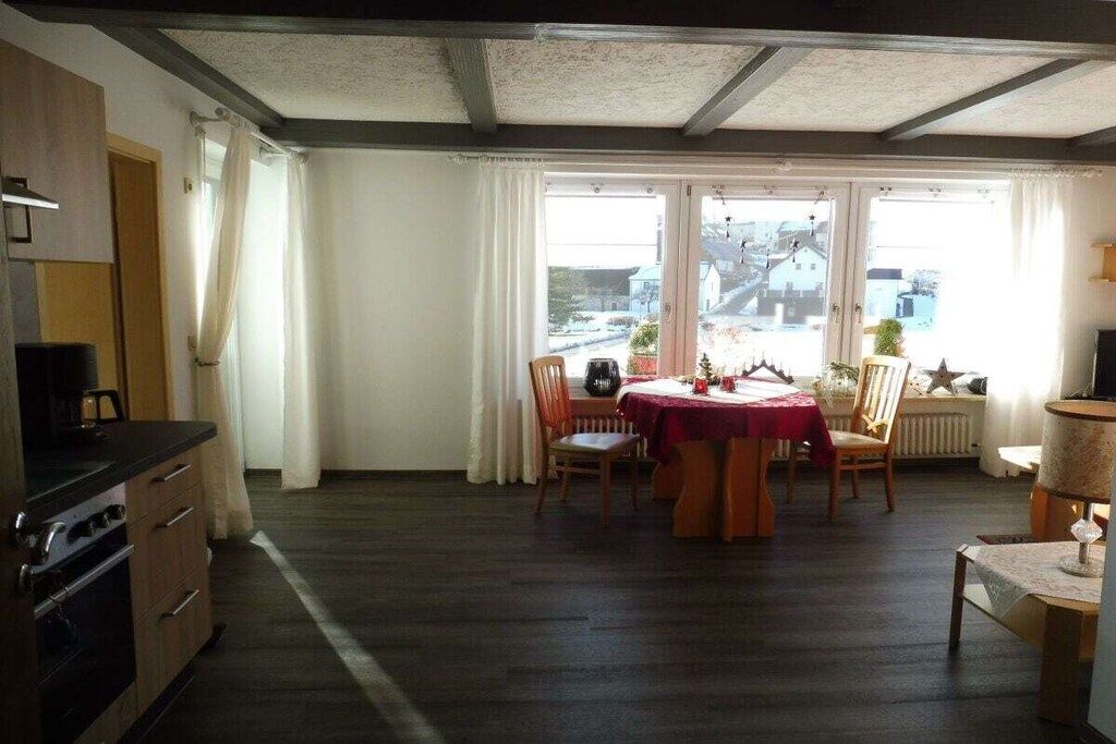 A dining area with a table set for four, a kitchen with a stove, and a view through large windows.