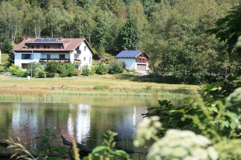 Ein Haus mit Solardach, einem Teich im Vordergrund und einer bewaldeten Hügelslandschaft im Hintergrund.