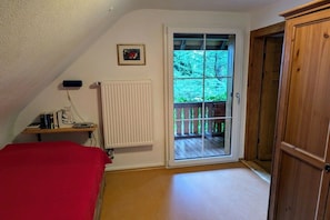 Een slaapkamer met een rode sprei, een radiator, een houten deur en een raam met uitzicht op een balkon.
