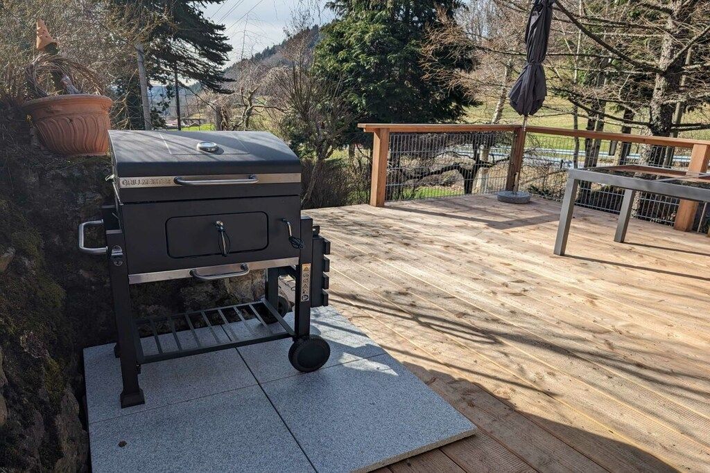 Ein Grill im Freien auf einer Holzterrasse, versehen mit einem Metallgeländer und einer angrenzenden Topfpflanze.