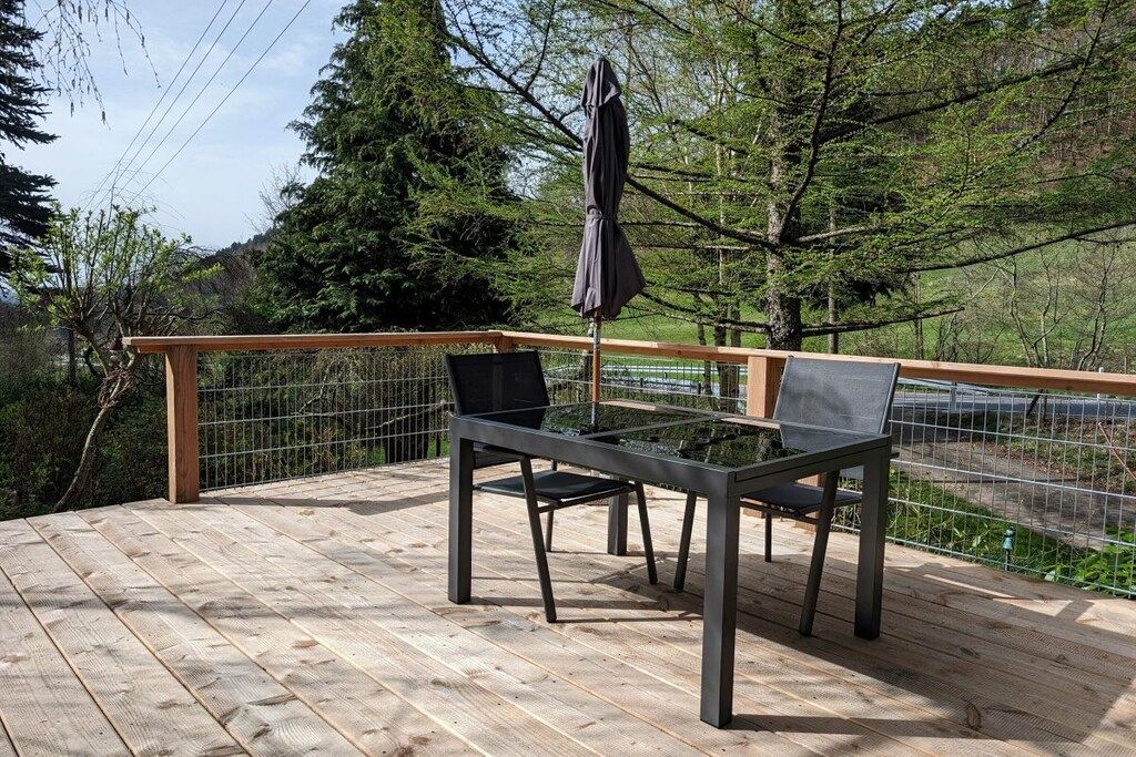 A wooden deck with a table and chairs, a metal railing, and a view of trees and hills.