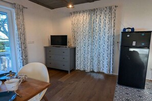 Une pièce de taille modeste avec un plancher en bois, une commode grise, un téléviseur, un réfrigérateur noir et une table à manger avec des chaises.