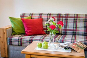 Ein kariertes Sofa mit einem roten und grünen Kissen, ein Holztisch mit einer Blumenvase, Äpfeln und einer Zeitschrift.
