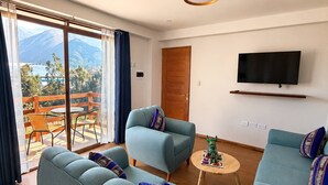 En moderne stue med sofa, lænestol og et lille bord. Et fjernsyn er monteret på væggen, og der er en skydedør, der fører til et udendørs område med udsigt til bjerge.