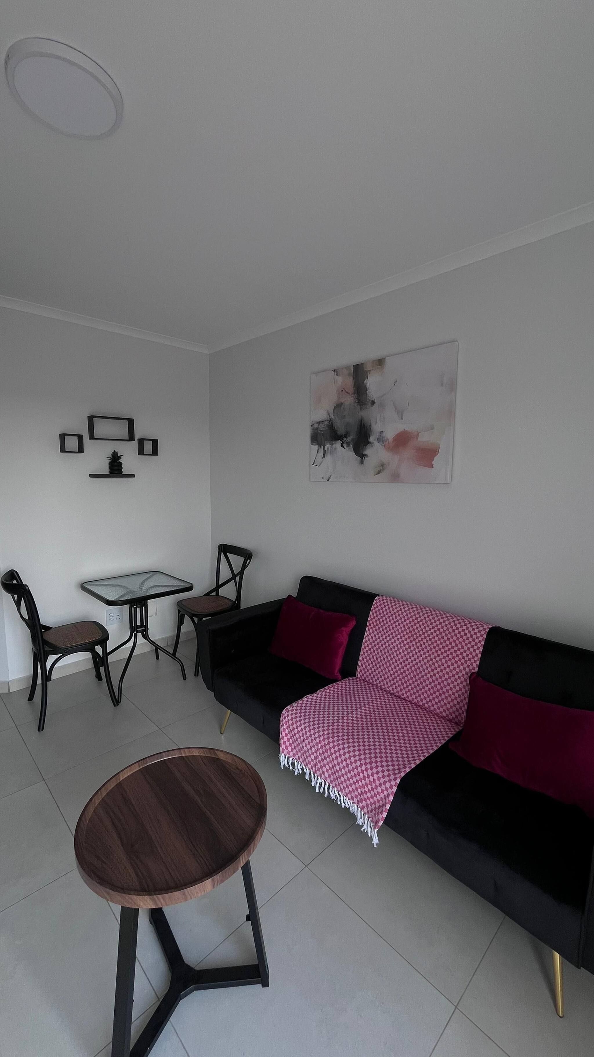 A modern living room with a black sofa, a round wooden coffee table, a small square table, two chairs, and a wall-mounted abstract painting.