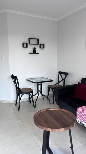 Ein Wohnzimmer mit einem runden Couchtisch, einer schwarzen Couch und zwei Sesseln. Es steht ein kleiner Tisch mit einer Glasplatte daneben, an der Wand befindet sich ein Blumentopf.