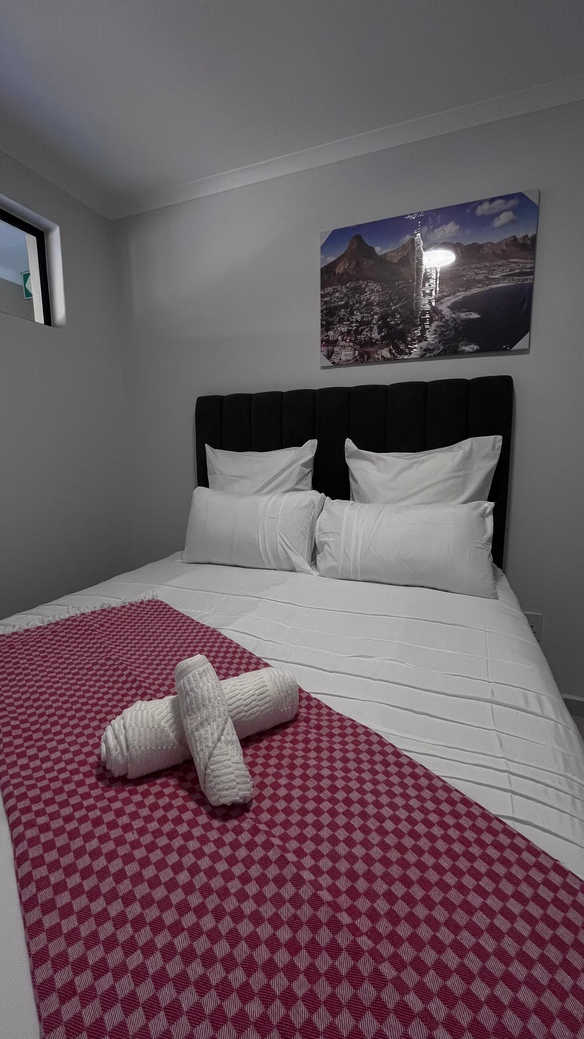 A hotel room with a bed, a red and white checkered blanket, white pillows, and a framed picture of a mountain landscape on the wall.