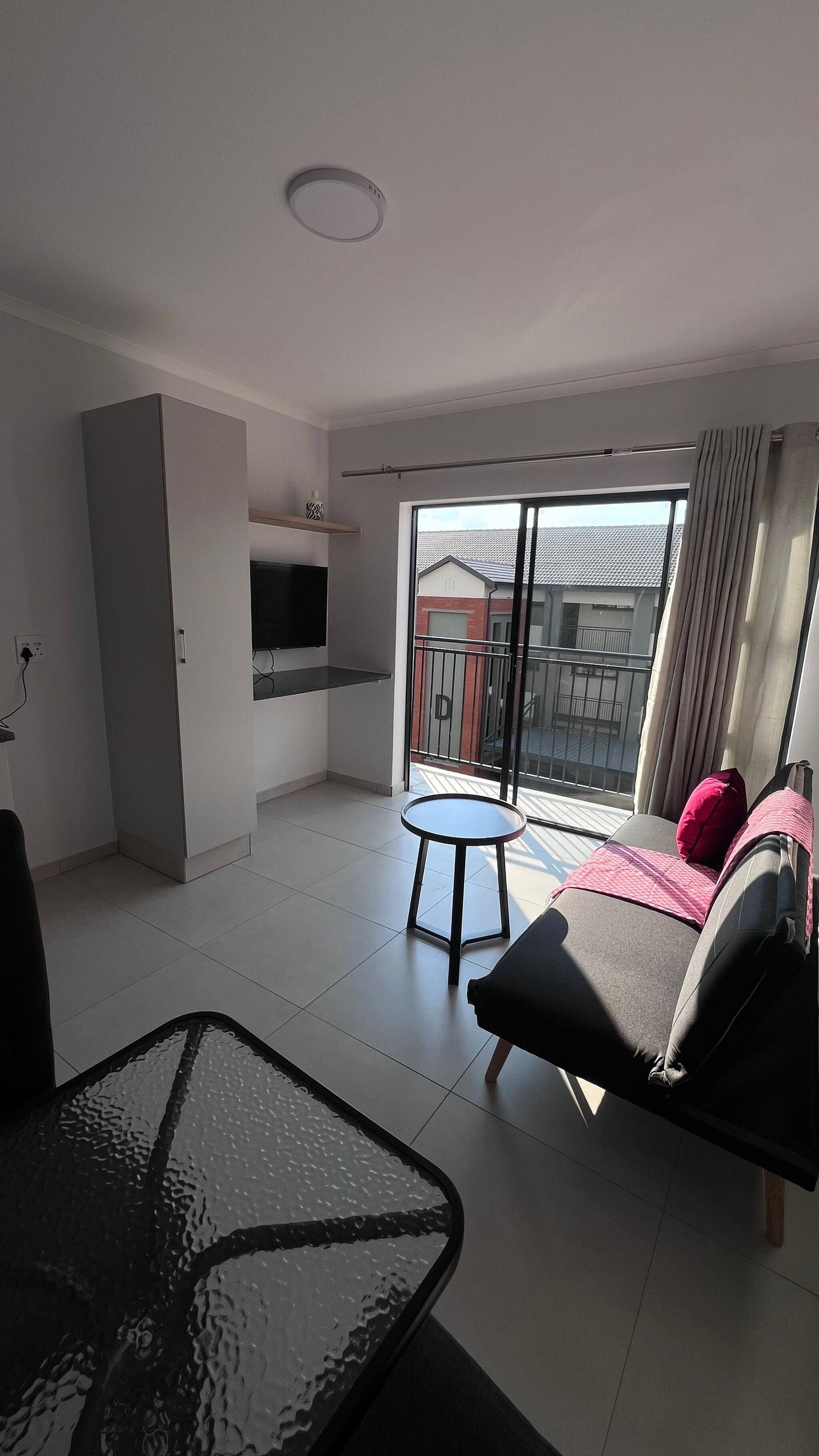 A modern living room with a glass dining table, a sofa with cushions, a TV mounted on the wall, and a balcony with a view of buildings.
