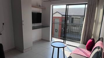 A modern living room with a glass dining table, a sofa with cushions, a TV mounted on the wall, and a balcony with a view of buildings.