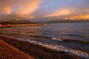 A waterfront view with a pier extending into calm waters, a beach in the foreground, and mountains in the background. - Oven bench directly on Lake Chiemsee (Chieming)